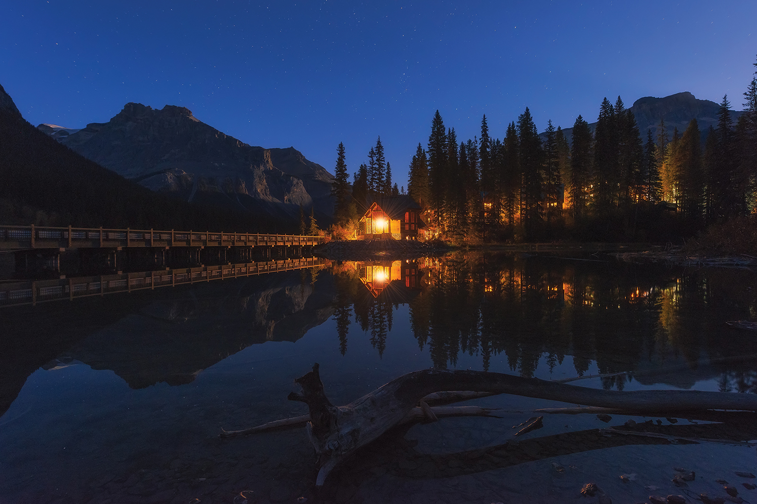 a small cabin on the lake with light on with woods and cabin reflected in water and stars seen in the sky