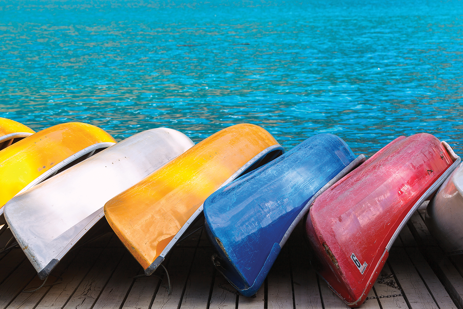 seven canoes including white, yellow, blue, and red, leaning on a dock by water