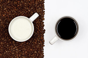 aerial view of two white mugs, one full of milk sitting on brown coffee beans and the other full of black coffee sitting on white
