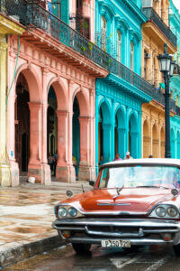 an old red taxi car parked on the street in front of pink, blue, and gold buildings