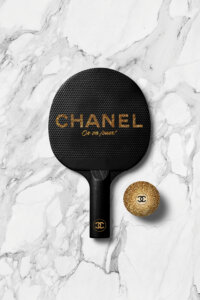 Black ping pong paddle with Chanel logo on it next to a gold Chanel ping pong ball on a white marble surface