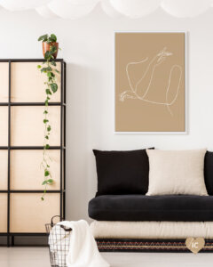 Tan colored graphic with figure-shaped white line drawing framed in white in a room with black and white sofa cushions and dresser with long ivy plant