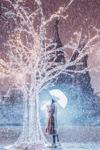a woman standing under a clear umbrella while looking at an all white tree with white lights on it while it's snowing