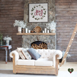 White shiplap sign that says "Joy" in red text with a wreath around it hanging on a stone fireplace with logs piled in it, white candles stacked on the mantle, and a white love seat near it