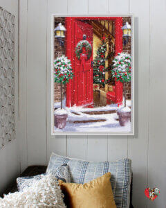 Art print of a red door with a Christmas wreath opening to the outdoors with snow hanging on a white shiplap wall next to a seat with yellow and white throw pillows