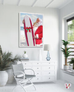 Print of Santa holding a red and white surfboard with a bow on it standing on the beach in a white coastal room with palm plants