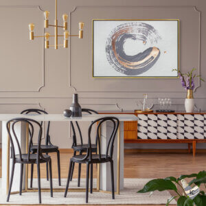 An abstract circular formation of brush strokes in a deep purple gray and metallic gold on a white background, framed in gold on a wall in a dining room setting