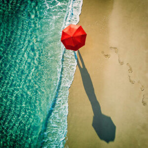 “Deep Water” by Ambra shows an aerial view of a person holding a red umbrella while walking down a sandy beach along the water’s edge and their elongated shadow.