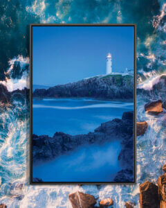 “Dusk I, Fanad Head Lighthouse, County Donegal, Ulster Province, Republic Of Ireland” by Gareth McCormack shows a blue sea surrounded by bank rocks under a blue sky with a lighthouse in the distance.