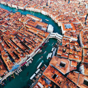“Grand Canal Aerial, Venice II” by Matteo Colombo shows an overhead view of buildings with orange rooftops surrounding a blue canal with boats in it.
