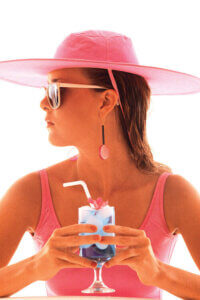 “1980s Young Woman In Bathing Suit Having A Drink” by Vintage Images shows the profile of a woman wearing a pink hat, pink bathing suit, and sunglasses while holding a drink.
