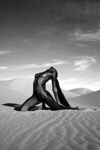 "Pyramid" by Gregory Prescott shows a black and white photo of a black woman with long hair sitting in a pyramid pose in a desert