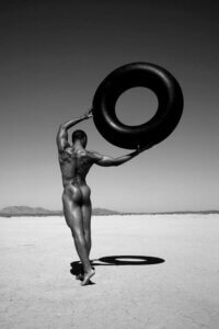 "Ramel With Tire" by Gregory Prescott shows a black and white photo of a black man walking through a desert holding a large black tire