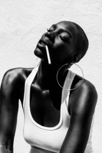 "Smoking" by Gregory Prescott shows a black and white photo of a black woman wearing a white tank top and large hoop earrings smoking a cigarette