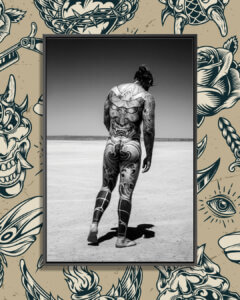 "Tattoo Guy" by Gregory Prescott shows a black and white photo of a nude man covered in tattoos walking through a desert