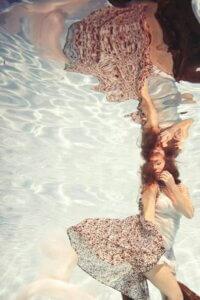 Ingenue by Lola Mitchell showcases a woman underwater wearing a burgundy and white dress with her reflection above her