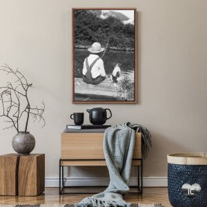 Minimal wood cabinet and black coffee set below a black and white framed photo of a boy in overalls facing a lake and fishing next to his dog