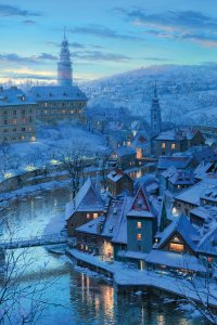 A snowcovered city near a river with several lights on in building windows.