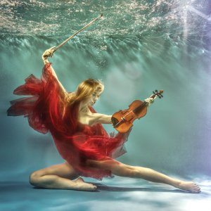 A woman underwater surrounded by a flowing red fabric while holding a violin.