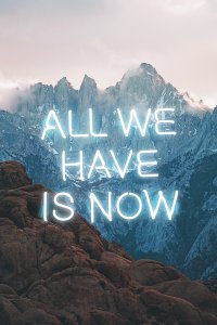 The phrase "All we have is now" in white neon letters in front of a mountainscape backdrop.