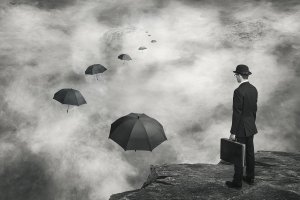 Man holding a suitcase standing on a cliff looking towards a path of floating umbrellas